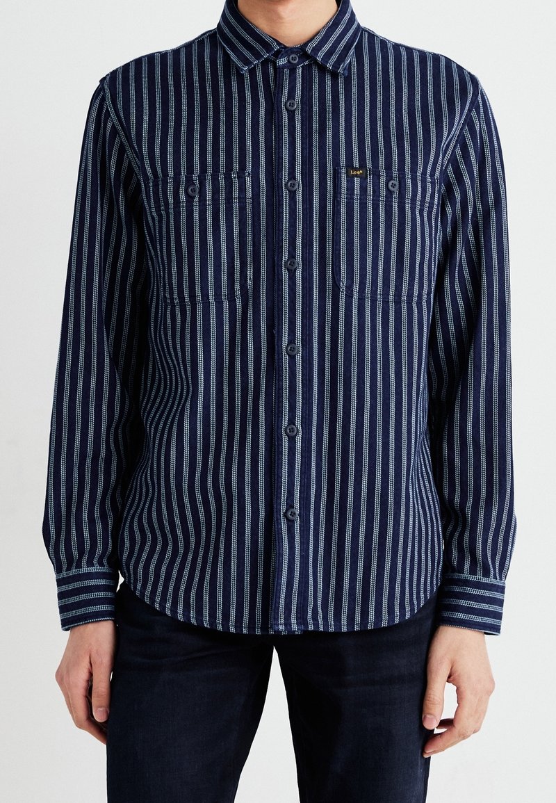 Man med en marinblå skjorta med knappar, vertikala vita ränder och två bröstfickor, kombinerad med mörka byxor.