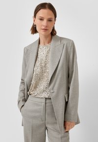 Vrouw in een lichtgrijze op maat gemaakte broekpak met een top versierd met pailletten, staand met één hand in de zak tegen een effen lichte achtergrond.