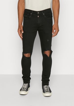 Homme portant un jean skinny noir déchiré avec des trous aux genoux et des baskets en toile noires et blanches, debout sur fond blanc.
