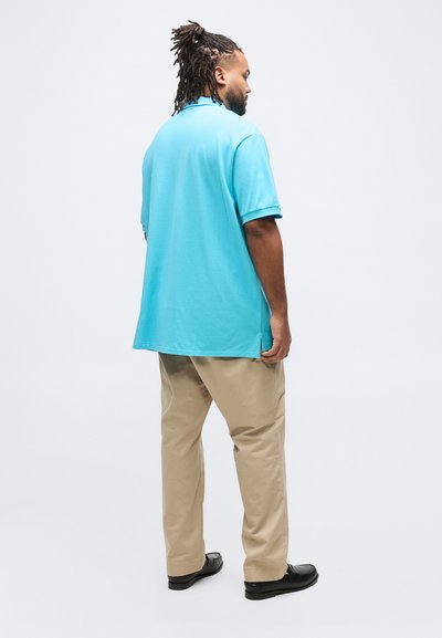 Άνδρας με κοτσίδες, φορώντας τιρκουάζ πόλο πουκάμισο, μπεζ παντελόνι και μαύρα παπούτσια, στέκεται σε λευκό φόντο, στραμμένος πλευρικά.