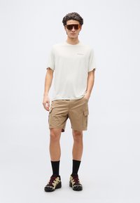 Νεαρός άνδρας που φοράει μπεζ cargo σορτς, ανοιχτό μπεζ μπλουζάκι Patagonia, μαύρες κάλτσες, μπεζ και μαύρα παπούτσια πεζοπορίας και αθλητικά γυαλιά ηλίου με κόκκινο φίλτρο.