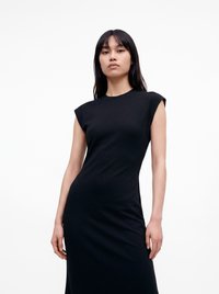 Jeune femme aux cheveux noirs raides portant une robe noire ajustée sans manches, debout devant un fond blanc uni.