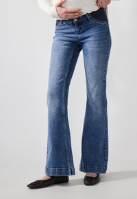 Jeans de denim azul acampanados con un lavado medio y un ligero desvanecimiento. Presentan un cierre de botón, bolsillos frontales y un ajuste entallado en la cintura.