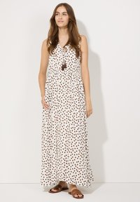 Robe maxi blanche à pois marron, décolleté en V, taille élastique et détail à franges. Le modèle porte des sandales plates marron, se tenant sur un fond blanc.