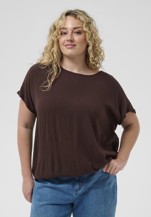 Blonde Frau mit lockigem Haar, die ein lockeres braunes Oberteil und blaue Jeans trägt, steht mit einer Hand in der Tasche vor schlichtem Hintergrund.