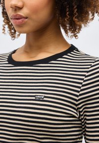 Donna che indossa una maglietta Levi's a righe orizzontali nere e beige con scollo a girocollo e capelli ricci parzialmente visibili su uno sfondo chiaro.