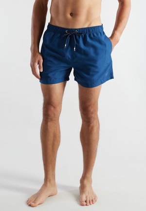 Oberkörperfreier Mensch, der blaue Badeshorts mit Kordelzug trägt und barfuß vor einem schlichten hellen Hintergrund steht.