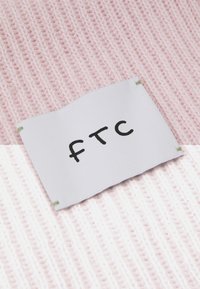 Pink knitted fabric with a white label featuring black text "FTC," attached with green stitching at the corners.