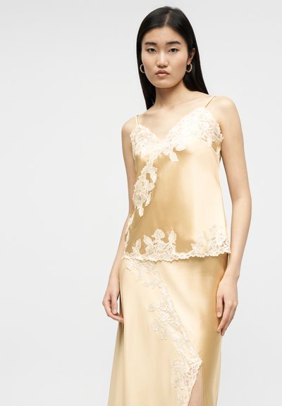 Γυναίκα που φοράει μπεζ σατέν καμιζόλ και ταιριαστή φούστα με διακοσμητικά κεντήματα λουλουδιών από δαντέλα, στέκεται μπροστά σε απλό φωτεινό φόντο.