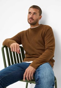 Bruine gebreide trui met ribbelstructuur, ronde hals, gedragen over een witte shirt, gecombineerd met blauwe jeans, zittend op een groene lattenstoel.