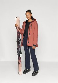 Χειμερινό μπουφάν σε σκουριά απόχρωση με μαύρες λεπτομέρειες, συνδυασμένο με μπλε σκούρο παντελόνι για σκι. Το μοντέλο κρατάει ροζ ανοιχτές σανίδες σκι με μαύρους δεσμούς. Μαύρα μπότες σκι.