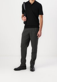 Man in een zwart poloshirt, grijs gestreepte broek, zwarte leren laarzen, die een zwarte schoudertas vasthoudt tegen een witte achtergrond.