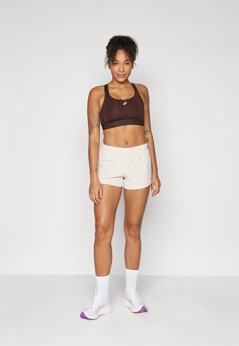 Brauner Sport-BH mit Racerback-Design, kombiniert mit hellbeigen Shorts mit elastischem Bund, abgerundet mit weißen Socken und Sportschuhen.