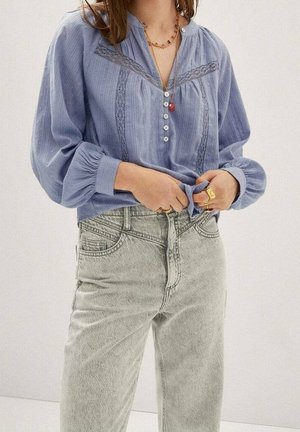 Kvinde iført en blå bluse med blonder og knapper, stoppet ned i højtaljede lysegrå jeans, accessoriseret med ringe og en halskæde.