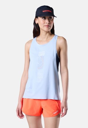 Frau trägt eine schwarze Rossignol-Kappe, ein hellblaues ärmelloses Oberteil mit grafischem Text und leuchtend orangefarbene Shorts vor einem schlichten Hintergrund.