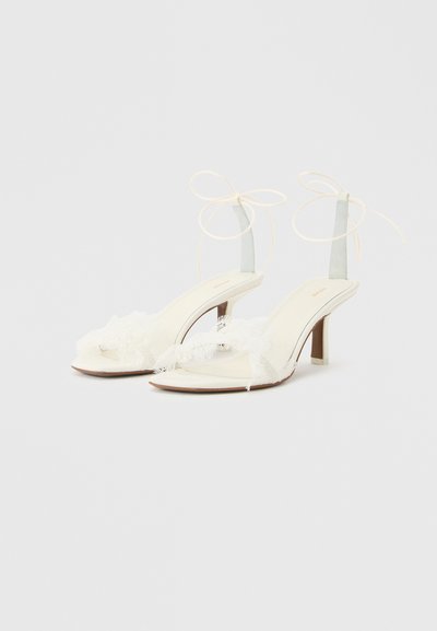 Λευκά σανδάλια με ψηλό τακούνι και λεπτά λουριά στον αστράγαλο δεμένα με φιόγκους, με λεπτή διακοσμητική φθαρμένη ύφανση στην περιοχή των δαχτύλων.