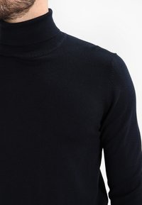 Maglione collo alto nero con colletto a costine, realizzato in un tessuto morbido e liscio, caratterizzato da un design aderente e dettagli di cucitura minimali.