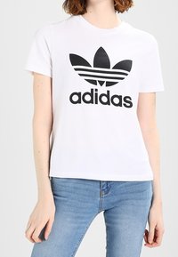 Camiseta blanca de algodón con un logo negro de Adidas y tres rayas negras encima. Combinada con vaqueros de denim azul claro.