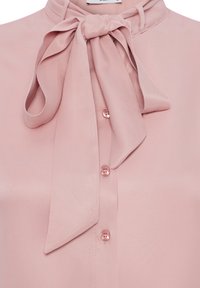 Blusa rosa chiaro con colletto a fiocco, bottoni frontali e un tessuto liscio e strutturato. Presenta un design classico e una lieve lucentezza.
