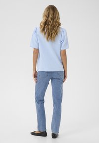 Top texturizado azul claro con mangas cortas abullonadas, combinado con jeans de mezclilla azul y flancos negros, visto desde la parte de atrás.