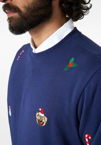 Mornarsko pulover s krožno ovratnikom vsebuje vezenine prazničnih motivov, vključno z mopsom v božični čeladi, snežakom in bomboni.