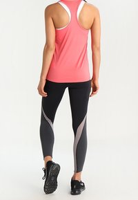 Vaaleanpunainen hihaton toppi, jossa on urheilullinen takatuki, yhdistetty mustiin legginsseihin, joissa on harmaita kulmakkaita yksityiskohtia ja teksturoituja mustia lenkkareita.