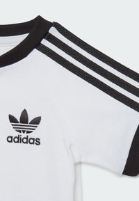 T-shirt bianco a maniche corte con colletto a costine nero e tre strisce nere sulle spalle. Presenta un logo ricamato nero sul petto.