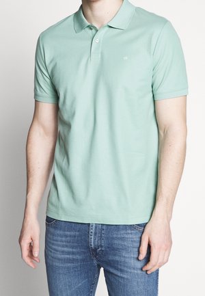 Hellgrünes Baumwoll-Poloshirt mit Kragen, kurzen Ärmeln und kleinem Logo auf der linken Brust, kombiniert mit blauen Jeans.