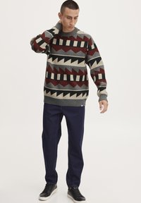 Knit sweater with geometric patterns in red, black, gray, and cream; crew neckline and ribbed cuffs. Paired with dark blue denim jeans.