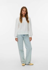 Femme portant un chemisier long à manches longues texturé blanc, un jean large bleu clair et des chaussures plates beiges, se tenant devant un fond uni.