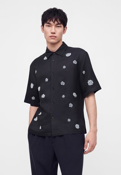 Jeune homme debout, portant une chemise noire à manches courtes avec des impressions florales blanches dispersées et un pantalon sombre.