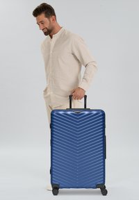 Man in een beige shirt en broek trekt een grote blauwe harde koffer met vier wielen op een witte achtergrond.