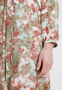 Robe florale aux teintes atténuées de verts, bleus et rouges. Tissu doux avec de longues manches froncées et un détail noué à la taille. Texture lisse.
