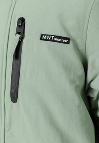 Veste vert clair avec une poche zippée, ornée d'un patch noir portant l'inscription "MNT depuis 1997." Texture lisse, design moderne.