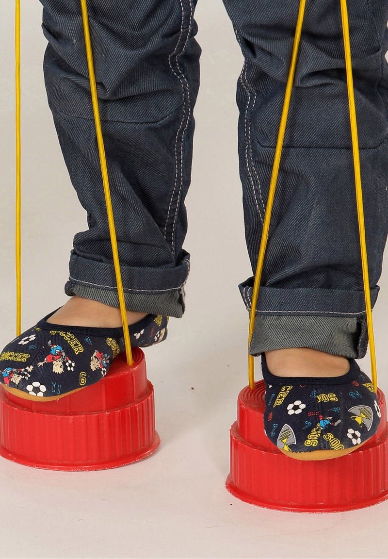 Enfant portant un jean foncé et des chaussons à motifs, debout sur des échasses jouets rouges avec des cordes jaunes sur un fond clair uni.