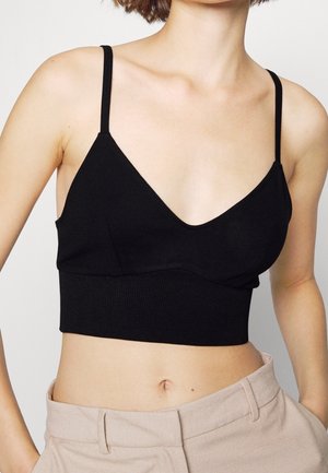 Kobieta w czarnym krótkim topie na cienkich ramiączkach i beżowych spodniach z wysokim stanem, odsłaniająca brzuch i obojczyki.