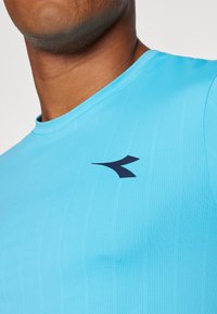 Himmelblaues Kurzarm-Sporthemd aus strukturiertem Stoff, mit geripptem Kragen und einem dunkelblauen Logo im linken Brustbereich.