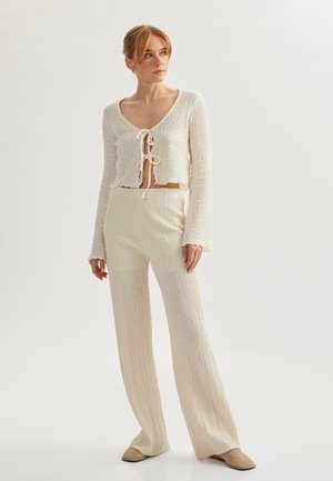 Strikoutfit bestående af en cropped, åben front, hvid top med bindedetalje og lange ærmer, parret med vidde ben, matchende teksturerede bukser.