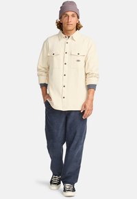 Crème-kleurig stoffen shirt met twee borstzakken, donkere knopen en grijze manchetten, gecombineerd met navy corduroy broeken en zwarte sneakers.