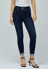 Los pantalones vaqueros ajustados de mezclilla azul oscuro presentan una cintura alta con dos botones dorados y puños enrollados. La tela tiene una textura suave con un ligero desgaste.