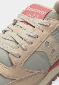 Κοντινό πλάνο του αθλητικού παπουτσιού Saucony Jazz που δείχνει ελαφρύ μπεζ σουέντ, λευκά υφασμάτινα κορδόνια, γκρίζο ύφασμα και ροζ λογότυπο στη γλώσσα.