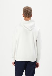 Sweatshirt à capuche blanche en coton texturé, avec une capuche spacieuse et des poignets côtelés, présenté de dos sur un fond uni.