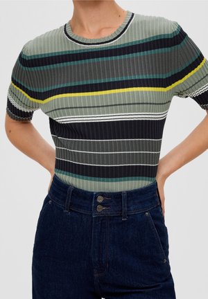 Person iført en ribstrikket kortærmet stribet top i grøn, sort, gul og hvid, stoppet ned i højttaljede mørkeblå jeans.