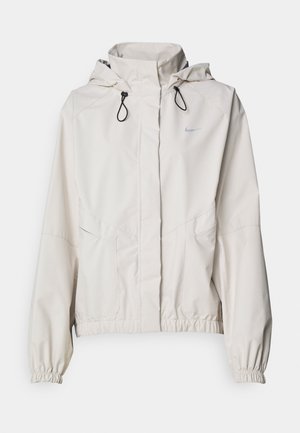 Lys beige zip-in-jakke med hette, elastiske mansjetter og to lommer foran. Har Nike-logoen i sølv på øvre venstre side.