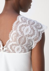 Haut blanc avec un corps en tissu côtelé et des manches délicates en dentelle ornées de motifs floraux et de bords en dentelle festonnée, fermé par des boutons à l'arrière.