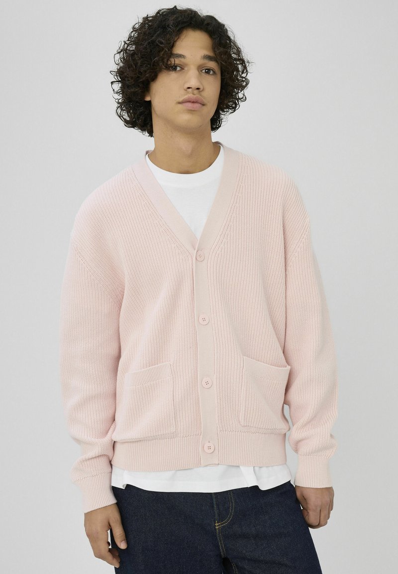 Cardigan rose en tricot avec un col en V, des boutons à l'avant et deux poches. Présente une texture côtelée et des manches longues, porté par-dessus une chemise blanche.