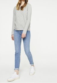 Nuori henkilö, joka käyttää vaaleanharmaata pitkähihaista paitaa, ylös käärittyjä sinisiä farkkuja ja valkoisia lenkkikenkiä, seisoo yksinkertaista valkoista taustaa vasten.