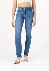 Femme portant un jean skinny bleu taille haute et des sandales à talons beiges, se tenant contre un fond blanc uni.