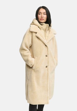 Beige nep-shearling jas met een brede kraag, zijzakken en knoopsluiting. Gelaagd over een lichtbeige pufferjack.