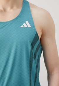 Türquoise sporditop, millel on tekstuuriline muster, valge logo vasakul õlal ja tumeroheline aktsenttriip külgedel.